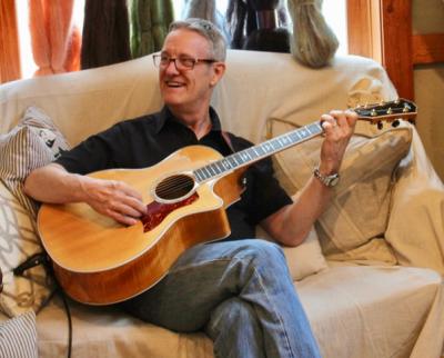 Ken Wheaton, seated on a couch and smiling, playing a guitar
